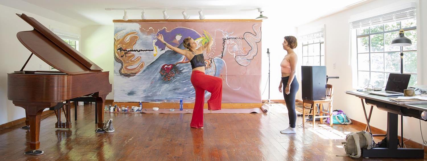 Two people standing in a room, facing each other. One person's leg and arms are raised. The other person is standing still.