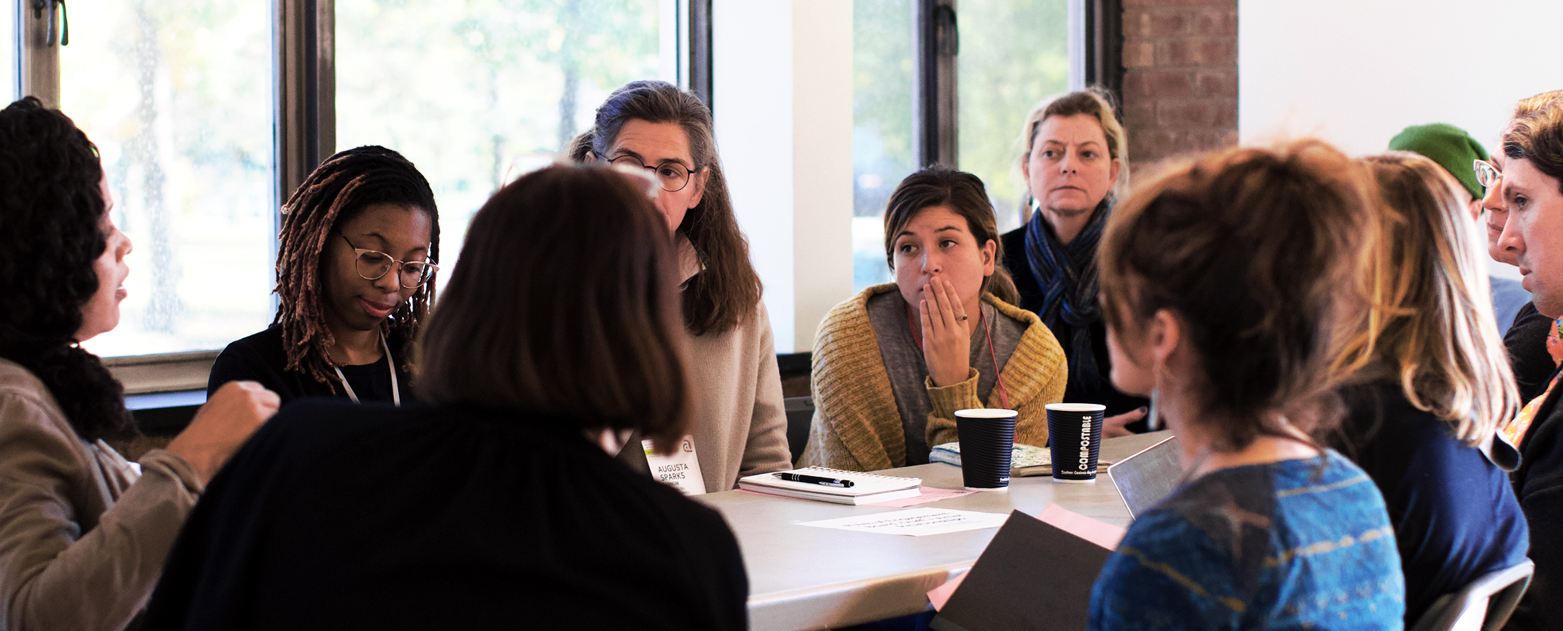 A group of people sitting around a table, listening to the person at the head of the table speak.