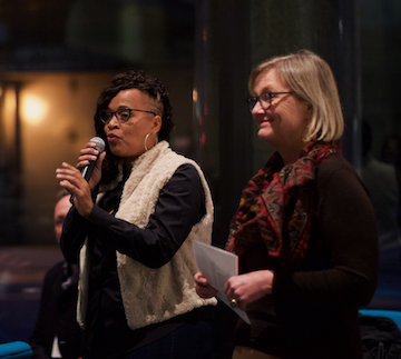 Two people standing next to each other. One is holding a microphone and gesturing to a crowd. The other is holding a piece of paper in both hands.