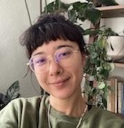Mid 30's non-binary Japanese American, gold glasses, dark brown hair, smiling, green shirt, plants in the background